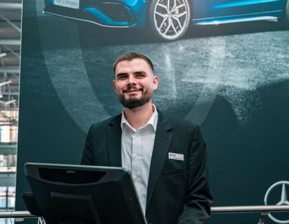 Dark-haired Jan Niklas Rakoci stands behind a reception desk on which there is a computer screen, a keyboard and a cup of coffee. In the background hangs a large dark poster with a blue car on it, a Mercedes star and the words ‘Made in Bremen’. He is wearing a white shirt and a black jacket.