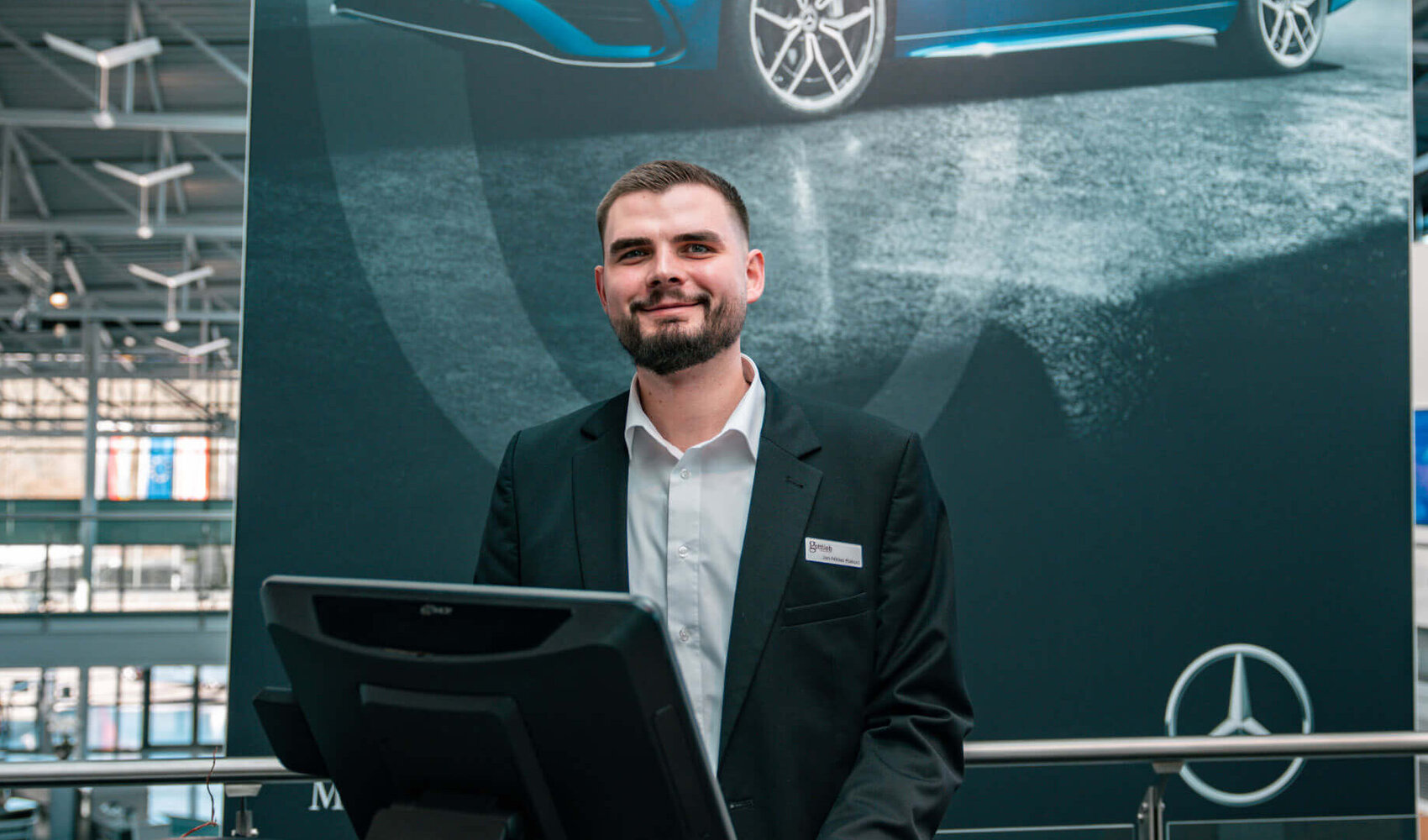 Der dunkelhaarige Jan Niklas Rakoci steht hinter einem Empfangstresen, auf welchem ein Computerbildschirm, eine Tastatur und ein Kaffee steht. Im Hintergrund hängt ein großes dunkles Plakat mit einem blauen Auto drauf, einem Mercedes Stern und der Aufschrift "Made in Bremen". Er trägt ein weißes Hemd und ein schwarzes Sakko. 