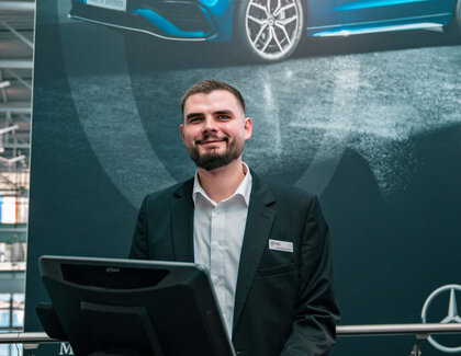 Der dunkelhaarige Jan Niklas Rakoci steht hinter einem Empfangstresen, auf welchem ein Computerbildschirm, eine Tastatur und ein Kaffee steht. Im Hintergrund hängt ein großes dunkles Plakat mit einem blauen Auto drauf, einem Mercedes Stern und der Aufschrift "Made in Bremen". Er trägt ein weißes Hemd und ein schwarzes Sakko. 