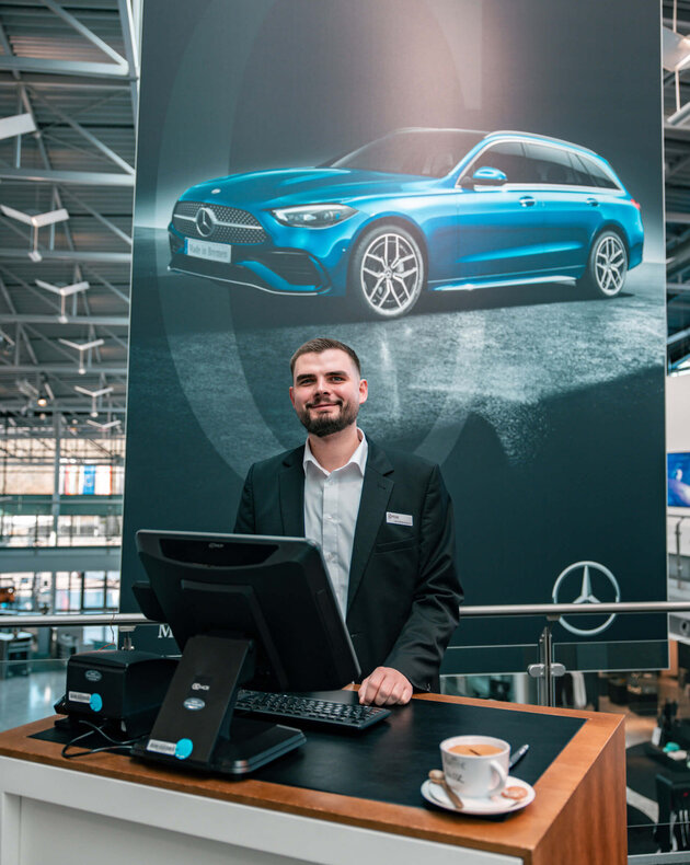 Der dunkelhaarige Jan Niklas Rakoci steht hinter einem Empfangstresen, auf welchem ein Computerbildschirm, eine Tastatur und ein Kaffee steht. Im Hintergrund hängt ein großes dunkles Plakat mit einem blauen Auto drauf, einem Mercedes Stern und der Aufschrift "Made in Bremen". Er trägt ein weißes Hemd und ein schwarzes Sakko. 