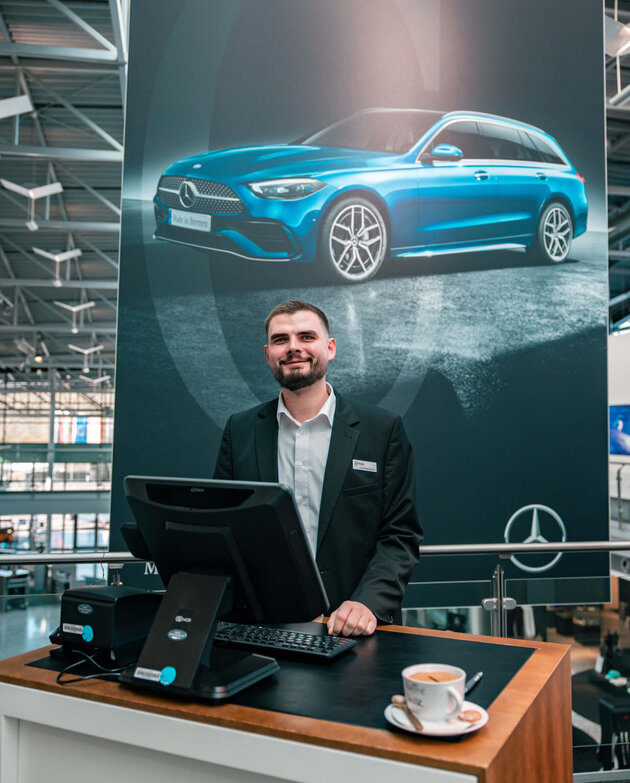 ATLANTIC success story – Deputy restaurant manager Jan Niklas Rakoci Dark-haired Jan Niklas Rakoci stands behind a reception desk on which there is a computer screen, a keyboard and a cup of coffee. In the background hangs a large dark poster with a blue car on it, a Mercedes star and the words ‘Made in Bremen’. He is wearing a white shirt and a black jacket.