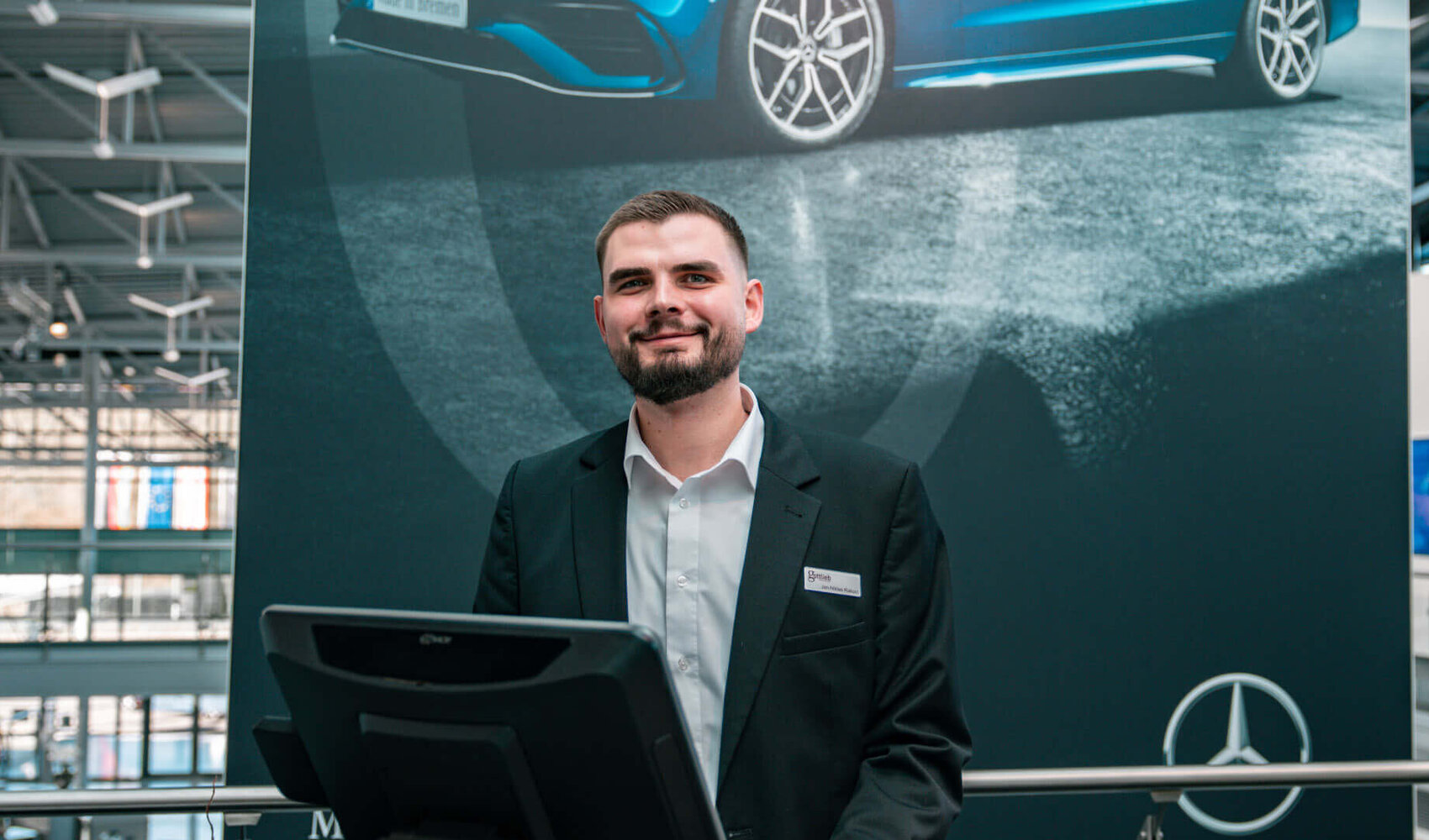 Dark-haired Jan Niklas Rakoci stands behind a reception desk on which there is a computer screen, a keyboard and a cup of coffee. In the background hangs a large dark poster with a blue car on it, a Mercedes star and the words ‘Made in Bremen’. He is wearing a white shirt and a black jacket.