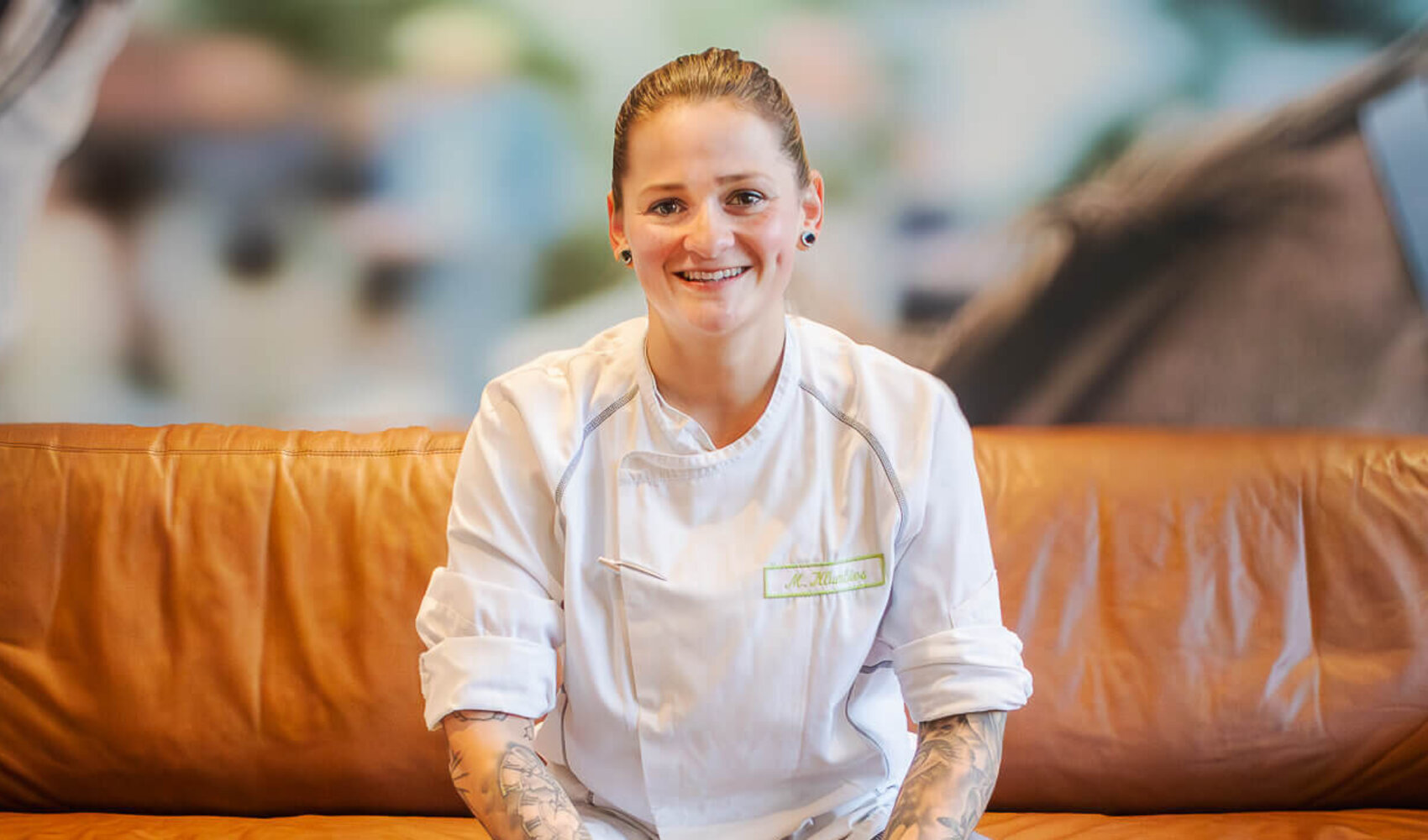 Die dunkelhaarige Madlin lächelt in Küchenuniform in die Kamera. Sie sitzt auf einem braunen Sofa. Vor ihr steht ein Kaffee Crema auf dem Tisch, daneben liegt ein Messer.  