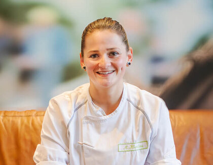 A coffee with Madlin Klumbies Dark-haired Madlin smiles at the camera in her kitchen uniform. She is sitting on a brown sofa. In front of her is a cup of coffee crema on the table, next to it is a knife.