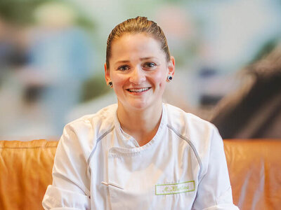 Dark-haired Madlin smiles at the camera in her kitchen uniform. She is sitting on a brown sofa. In front of her is a cup of coffee crema on the table, next to it is a knife.  