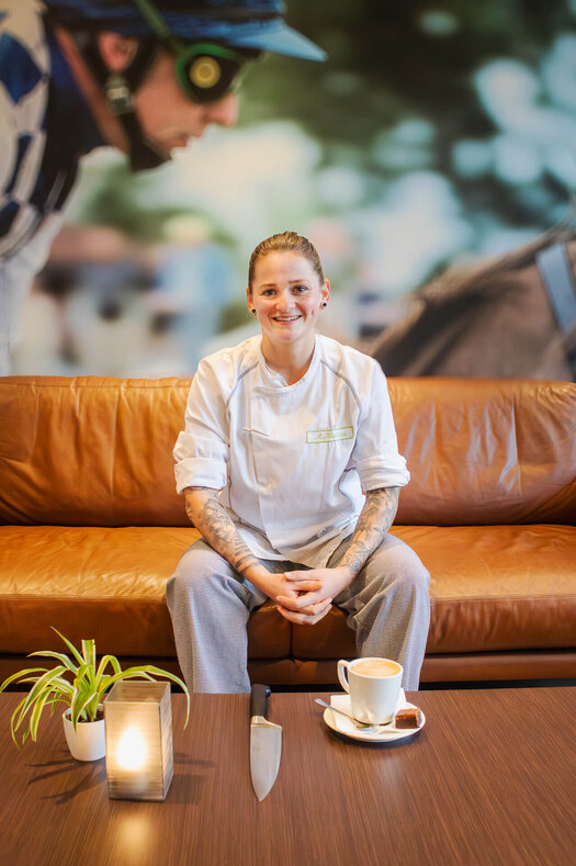 Dark-haired Madlin smiles at the camera in her kitchen uniform. She is sitting on a brown sofa. In front of her is a cup of coffee crema on the table, next to it is a knife.  