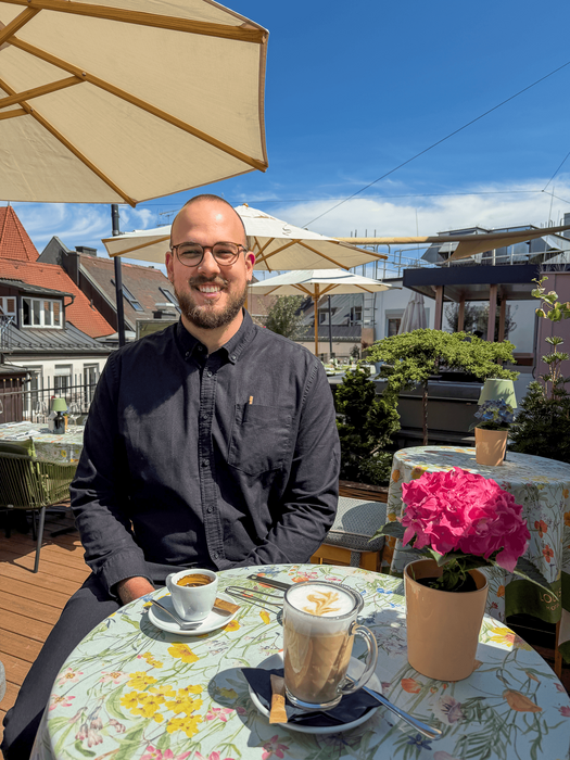 Igor Pocrnja trägt einen dunklen Bart und eine Brille. Er sitzt lächelnd an einem runden Tisch auf der Dachterrasse. Auf dem Tisch vor ihm stehen ein Espresso, ein Latte Macchiato und eine Topfblume. 