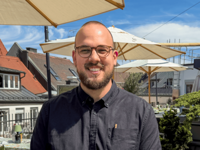 Igor Pocrnja trägt einen dunklen Bart und eine Brille. Er sitzt lächelnd an einem runden Tisch auf der Dachterrasse. Auf dem Tisch vor ihm stehen ein Espresso, ein Latte Macchiato und eine Topfblume. 
