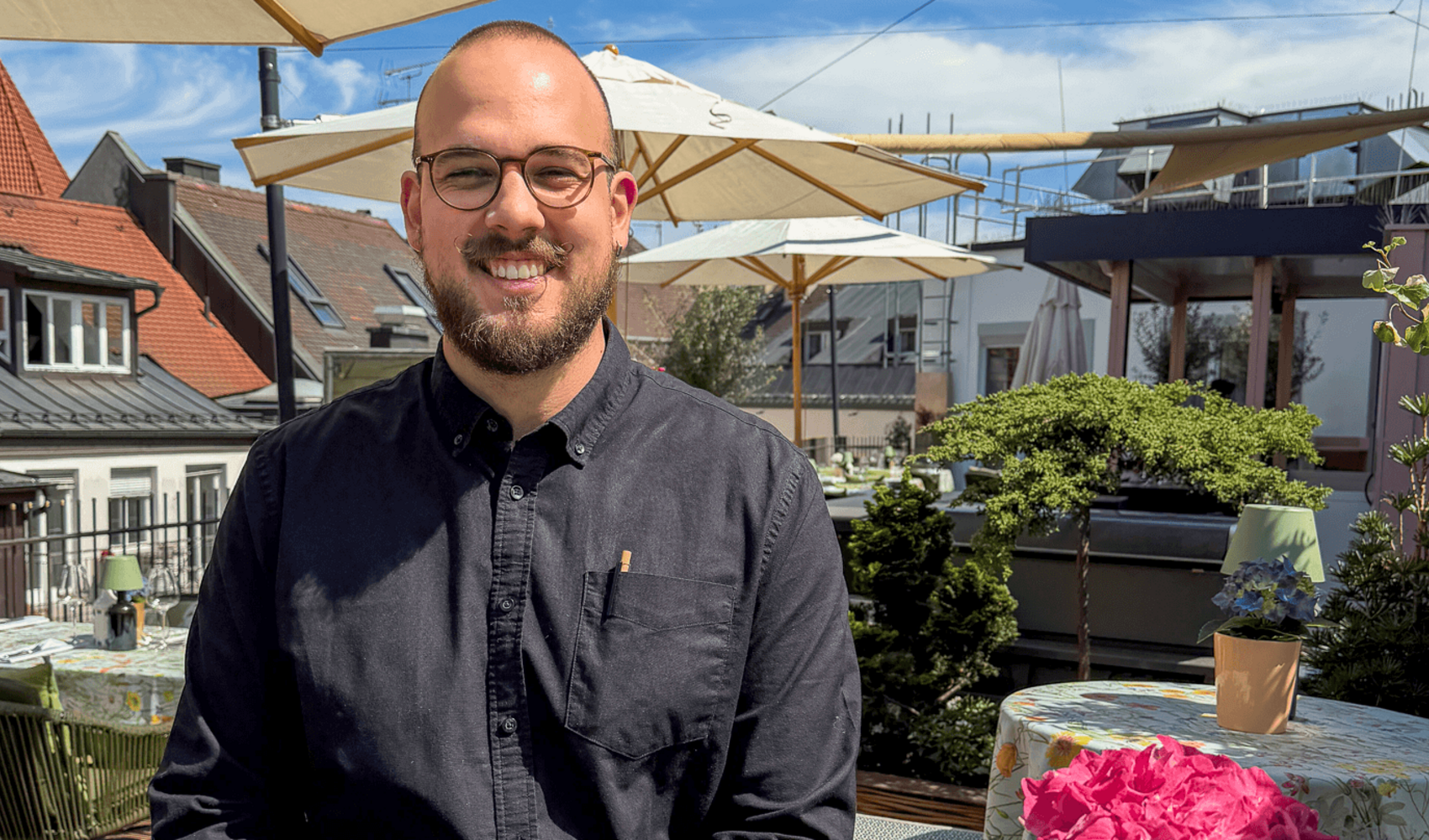 Auf einen Kaffee mit Igor Pocrnja (Bartender, LOUIS Hotel) Igor Pocrnja trägt einen dunklen Bart und eine Brille. Er sitzt lächelnd an einem runden Tisch auf der Dachterrasse. Auf dem Tisch vor ihm stehen ein Espresso, ein Latte Macchiato und eine Topfblume.
