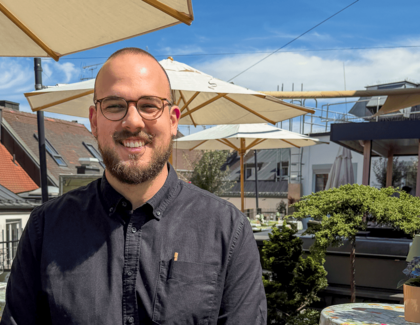 Igor Pocrnja trägt einen dunklen Bart und eine Brille. Er sitzt lächelnd an einem runden Tisch auf der Dachterrasse. Auf dem Tisch vor ihm stehen ein Espresso, ein Latte Macchiato und eine Topfblume. 