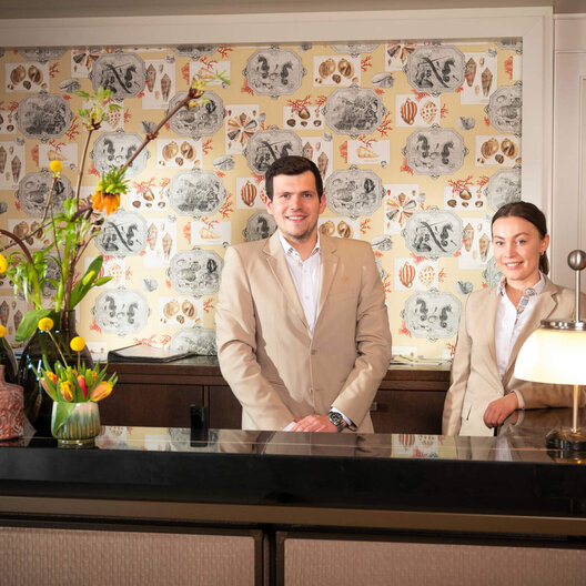 Mitarbeitende an der Rezeption des Severin*s Resort & Spa Ein männlicher und eine weibliche Mitarbeiter*in beigen Anzügen stehen an der Rezeption des Severin*s Resort & Spa und lächeln in die Kamera.