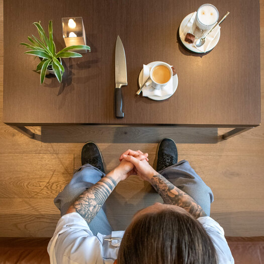 Photographed from above, dark-haired Madlin can be seen sitting at a brown wooden table. Her hands are folded in her lap. A coffee crema and a knife are on the table in front of her.  