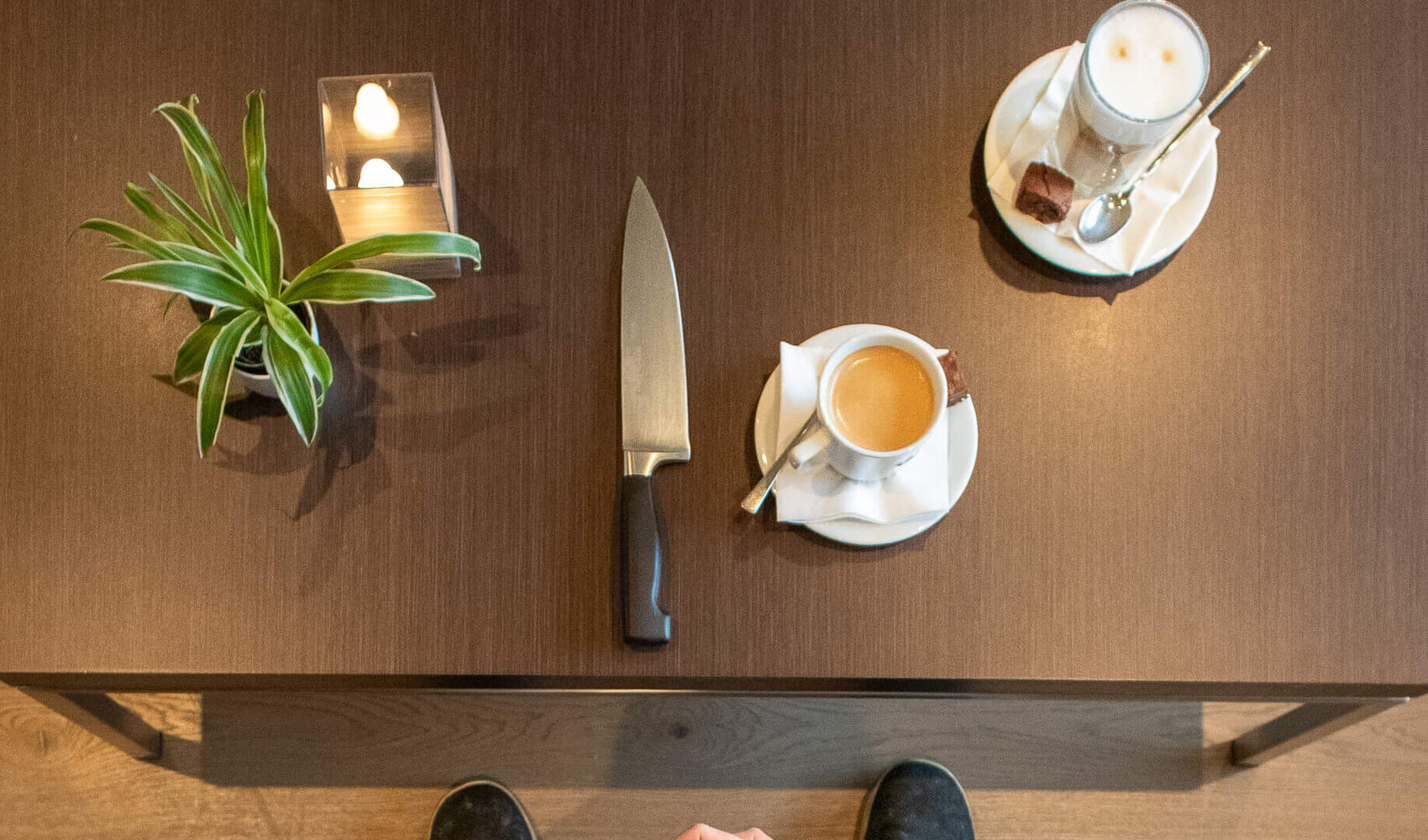 Photographed from above, dark-haired Madlin can be seen sitting at a brown wooden table. Her hands are folded in her lap. A coffee crema and a knife are on the table in front of her.  