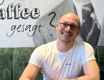 Der Operation Manager Christian Preißler sitzt lächelnd an einem Café-Tisch. Er trägt eine Brille und ein weißes Shirt mit dem Logo "unique Bremen". Auf dem Tisch vor ihm stehen zwei Cappuccini. Im Hintergrund ist "Hat hier jemand Kaffee gesagt" zu lesen.