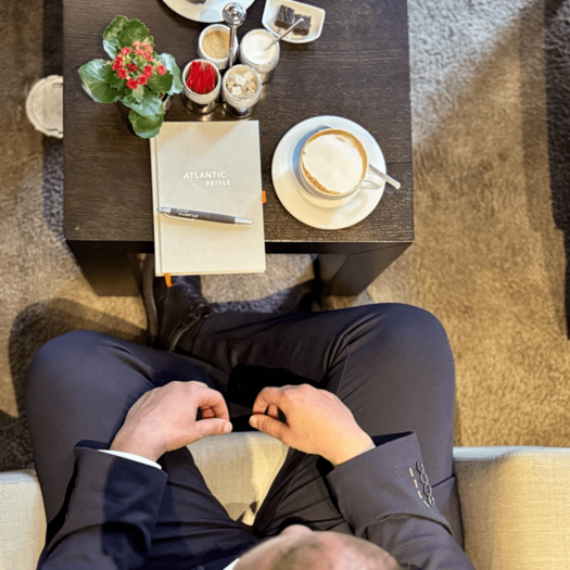 Auf einen Kaffee mit Front Office Manager Amr Ahmed Das Bild ist von oben fotografiert. Der dunkelhaarige Amr Ahmed sitzt auf einem beigen Sessel. Auf dem Tisch vor ihm steht ein Kaffee, ein Notizbuch und eine Blume.