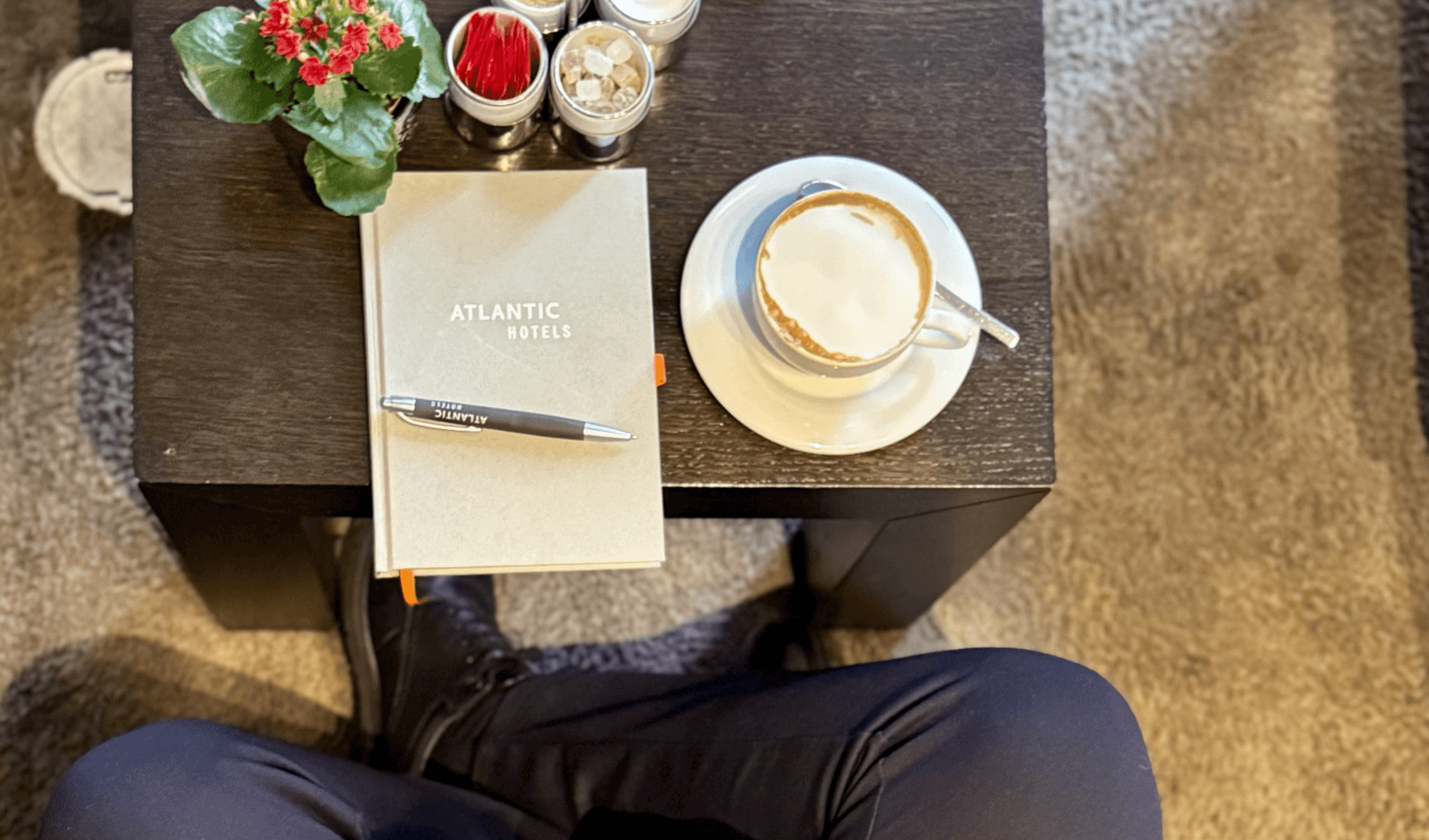 Auf einen Kaffee mit Front Office Manager Amr Ahmed Das Bild ist von oben fotografiert. Der dunkelhaarige Amr Ahmed sitzt auf einem beigen Sessel. Auf dem Tisch vor ihm steht ein Kaffee, ein Notizbuch und eine Blume.