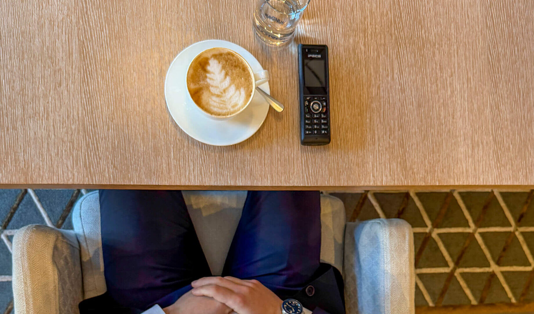 Von oben fotografiert ist der dunkelhaarige Cem Eryamansitzt glücklich an einem Holztisch. Auf dem Tisch vor ihm stehen zwei Cappuccini sowie zwei Wassergläser. Ein mobiles Telefon liegt ebenfalls auf dem Tisch. Im Hintergrund sind ein Holzschrank und eine Weltkarte zu sehen.