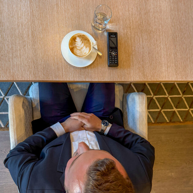 Von oben fotografiert ist der dunkelhaarige Cem Eryamansitzt glücklich an einem Holztisch. Auf dem Tisch vor ihm stehen zwei Cappuccini sowie zwei Wassergläser. Ein mobiles Telefon liegt ebenfalls auf dem Tisch. Im Hintergrund sind ein Holzschrank und eine Weltkarte zu sehen.