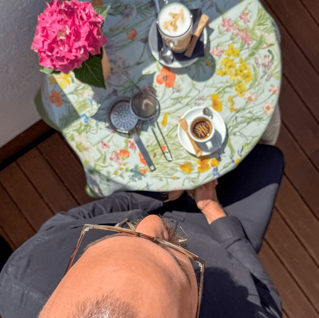 Von oben fotografiert ist Igor Pocrnja zu sehen, wie er an einem runden Tisch sitzt. Auf dem Tisch vor ihm stehen ein Espresso, ein Latte Macchiato und eine Topfblume. 