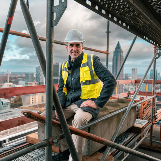 Ein Mann mit Bauhelm und Neonweste sitzt auf einem Baugerüst, im Hintergrund ist die Skyline von Frankfurt zu sehen.