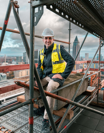 Ein Mann mit Bauhelm und Neonweste sitzt auf einem Baugerüst, im Hintergrund ist die Skyline von Frankfurt zu sehen.