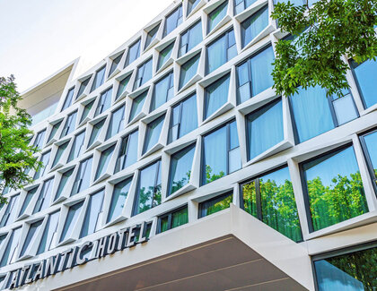 ATLANTIC Hotel Münster Außenansicht Eine Außenansicht vom ATLANTIC Hotel Münster. Über dem Eingang steht "ATLANTIC Hotel". Das Hotel hat sehr viele Fenster, welche von weißen Rahmen umgeben sind. Vor dem Hotel stehen grüne Bäume.