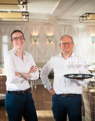 Service-Mitarbeitende des Severin*s Resort & Spa Zwei Mitarbeitende im Restaurant, beide in dunklen Hosen und hellen Hemden; der Kollege trägt ein Tablett mit Gläsern und beide lächeln in die Kamera.