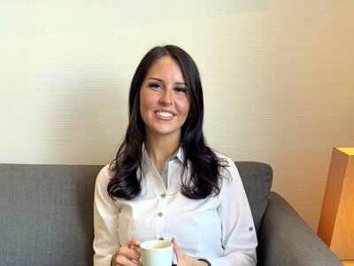 Die dunkelhaarige Paula Schulze sitzt an einem dunklen, kleinen Holztisch  und schaut in die Kamera. In ihren Händen hält sie eine Tasse Kaffee. Vor ihr auf dem Tisch steht ein weiterer Kaffee und kleines Gebäck. Sie trägt eine weiße Bluse und eine dunkle Hose.