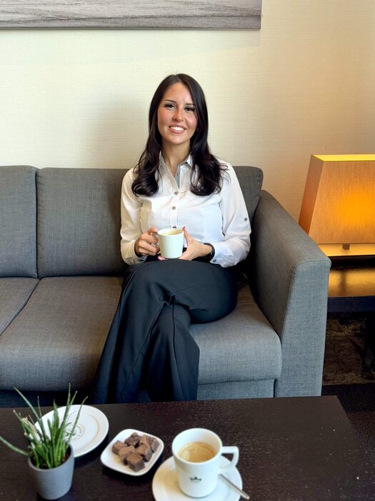 Die dunkelhaarige Paula Schulze sitzt an einem dunklen, kleinen Holztisch  und schaut in die Kamera. In ihren Händen hält sie eine Tasse Kaffee. Vor ihr auf dem Tisch steht ein weiterer Kaffee und kleines Gebäck. Sie trägt eine weiße Bluse und eine dunkle Hose.