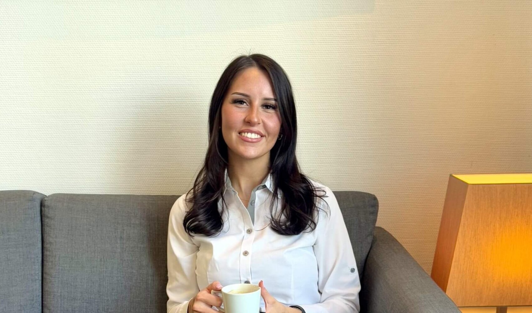 Die dunkelhaarige Paula Schulze sitzt an einem dunklen, kleinen Holztisch  und schaut in die Kamera. In ihren Händen hält sie eine Tasse Kaffee. Vor ihr auf dem Tisch steht ein weiterer Kaffee und kleines Gebäck. Sie trägt eine weiße Bluse und eine dunkle Hose.
