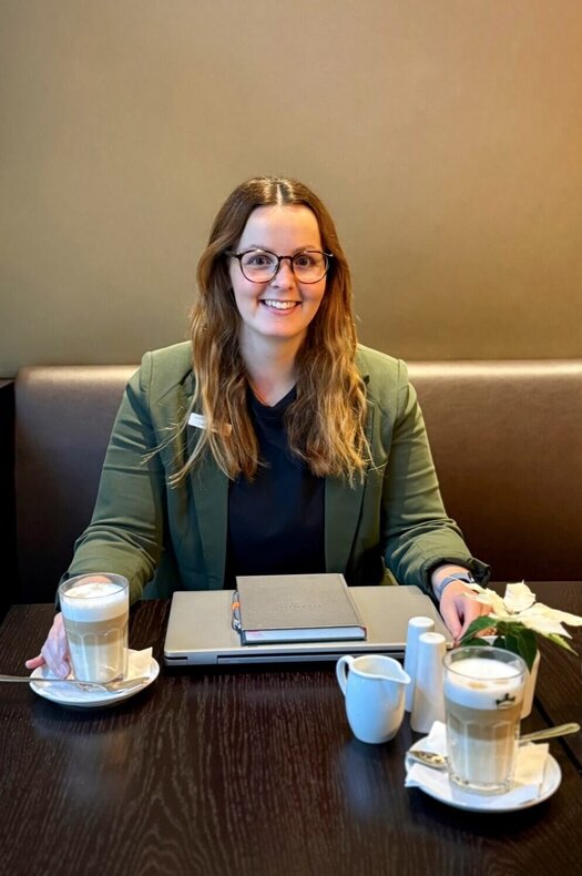 A coffee with Laura Sophie Keller Brown-haired Laura is sitting on a bench with a table in front of her. On the table are two coffees, a laptop, a notebook, a flower, and milk. She is wearing a green blazer and a dark top.