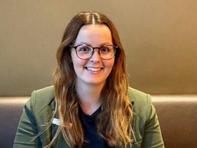 Brown-haired Laura is sitting on a bench with a table in front of her. On the table are two coffees, a laptop, a notebook, a flower, and milk. She is wearing a green blazer and a dark top.