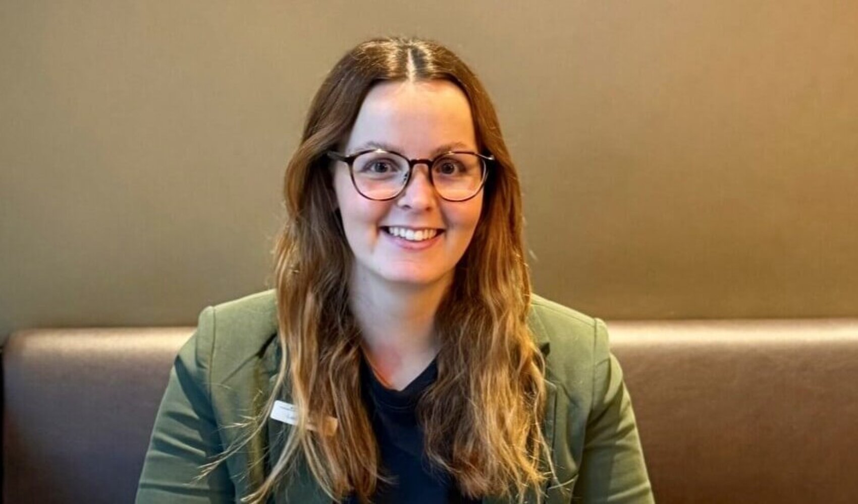 Die braunhaarige Laura sitzt auf einer Bank und vor ihr steht ein Tisch. Auf dem Tisch stehen 2 Kaffee, ein Laptop, ein Notizbuch, eine Blume und Milch. Sie trägt einen grünen Blazer und ein dunkles Oberteil.