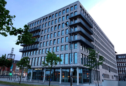 Eine Außenansicht vom ATLANTIC Hotel Kiel. Vor dem Hotel ist eine Straße und ein Rasenstreifen mit vier Bäumen. 