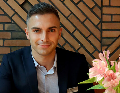 The dark-haired revenue manager Hardy Pape is sitting at a restaurant table with a stone wall in the background. He has one arm on the table. In front of him is a calculator. There are also two cappuccinos, a sugar glass and a pink flower on the table.