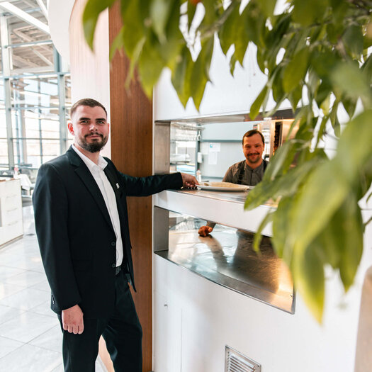 Der dunkelhaarige Jan Niklas Rakoci steht vor der Ausgabe aus der Küche und hält mit einer Hand einen Teller fest. Im Hintergrund in der Küche sieht man eine männliche Person, die in die Kamera guckt. Jan Niklas Rakoci trägt ein weißes Hemd, eine schwarze Hose und ein schwarzes Sakko. 
