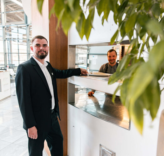 ATLANTIC success story – Deputy restaurant manager Jan Niklas Rakoci Dark-haired Jan Niklas Rakoci stands in front of the kitchen counter, holding a plate in one hand. In the background, a man can be seen looking at the camera. Jan Niklas Rakoci is wearing a white shirt, black trousers and a black jacket.