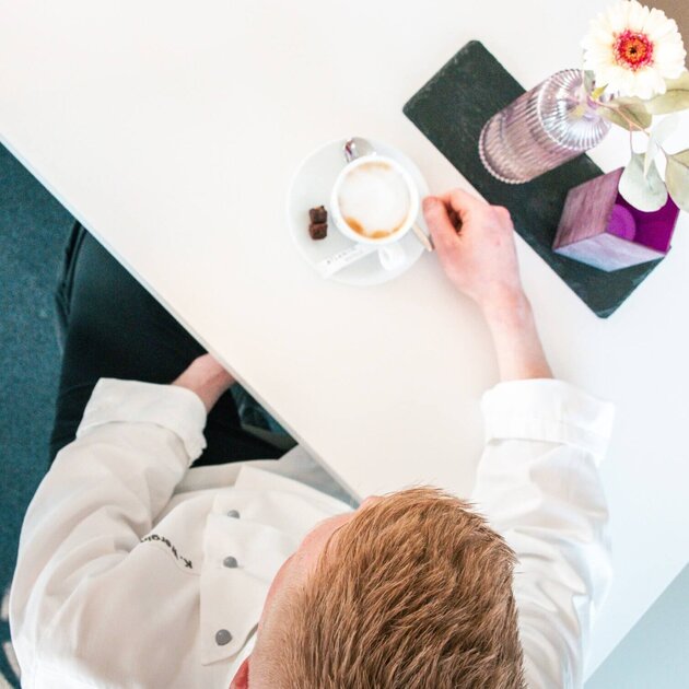 Kevin Wergin ist von oben am Tisch sitzend zu sehen. Es stehen ein Kaffee und Blumen auf dem Tisch.
