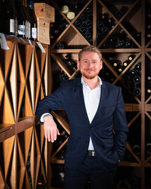 Severin*s Resort & Spa | Mitarbeiter im Weinkeller Ein junger Mann in einem weißen Hemd und einem dunklen Anzug steht in einem Weinkeller. Er lehnt mit einem Arm lässig an den Regalen.