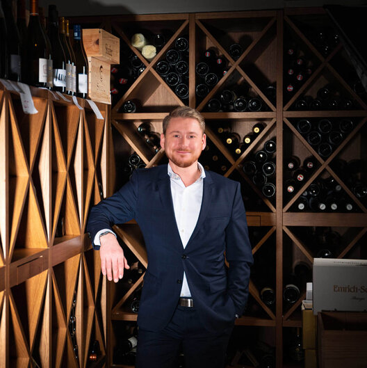 Severin*s Resort & Spa | Mitarbeiter im Weinkeller Ein junger Mann in einem weißen Hemd und einem dunklen Anzug steht in einem Weinkeller. Er lehnt mit einem Arm lässig an den Regalen.