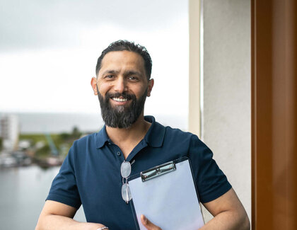 Der dunkelhaarige Bilal Madi steht auf einem Balkon und hält ein Klemmbrett in der Hand. Er trägt ein dunkelblaues Oberteil und eine dunkle Hose. Die Aussicht von dem Balkon ist aufs Wasser. 