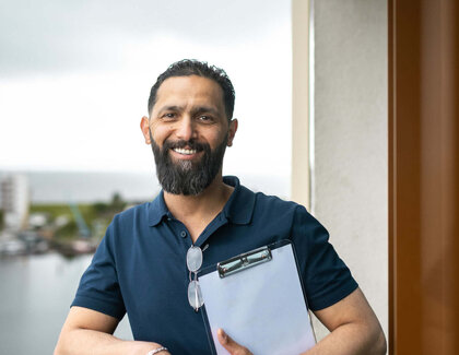 ATLANTIC success story - Deputy Housekeeping Manager Bilal Madi Dark-haired Bilal Madi is standing on a balcony, holding a clipboard in his hand. He is wearing a dark blue top and dark trousers. The view from the balcony is of the water.