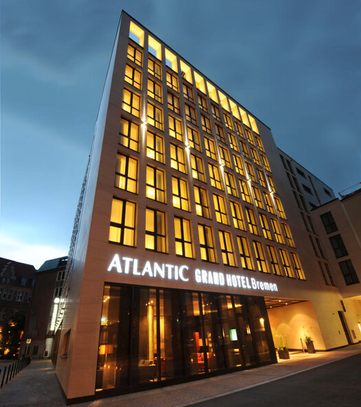 Eine Außenansicht vom ATLANTIC Grand Hotel Bremen bei Dämmerung. Alle Fenster der vorderen Hotelfront sind beleuchtet und über dem Eingang steht "ATLANTIC Grand Hotel Bremen".