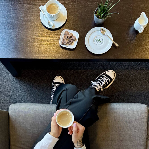 Von oben fotografiert ist die dunkelhaarige Paula Schulze zu sehen wie er an einem dunklen Holztisch sitzt. In den Händen hält sie einen Kaffee und auf dem Tisch vor ihr steht ein Kaffee, Gebäck und eine Pflanze. 