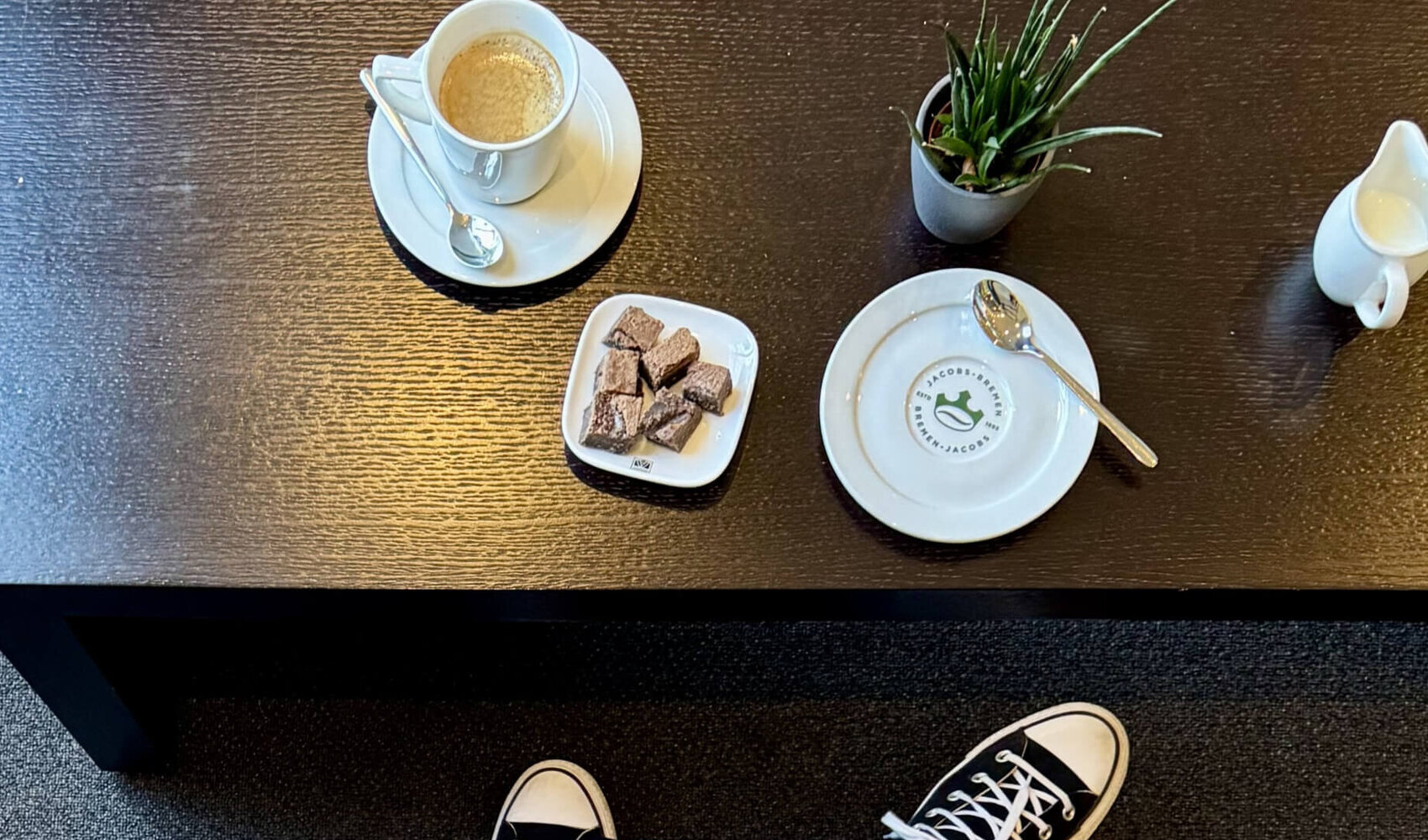 Von oben fotografiert ist die dunkelhaarige Paula Schulze zu sehen wie er an einem dunklen Holztisch sitzt. In den Händen hält sie einen Kaffee und auf dem Tisch vor ihr steht ein Kaffee, Gebäck und eine Pflanze. 
