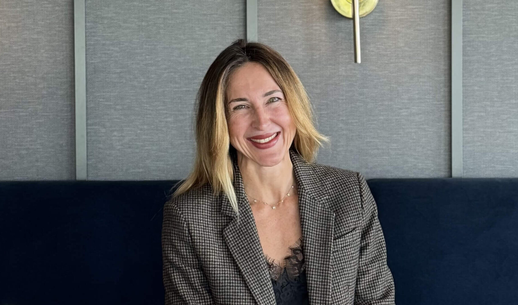 Auf einen Kaffee mit Yasmina Schaller (Rooms Division Managerin, ATLANTIC Hotel Münster) Die blonde Yasmina Schaller (Rooms Division Managerin) sitzt an einem dunklen Tisch in der Bar. Auf dem Tisch vor ihr stehen zwei Cappuccino und ein Telefon. Yasmina trägt einen Blazer.