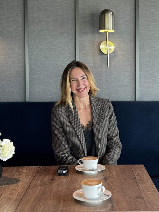 Die blonde Yasmina Schaller (Rooms Division Managerin) sitzt an einem dunklen Tisch in der Bar. Auf dem Tisch vor ihr stehen zwei Cappuccino und ein Telefon. Yasmina trägt einen Blazer.