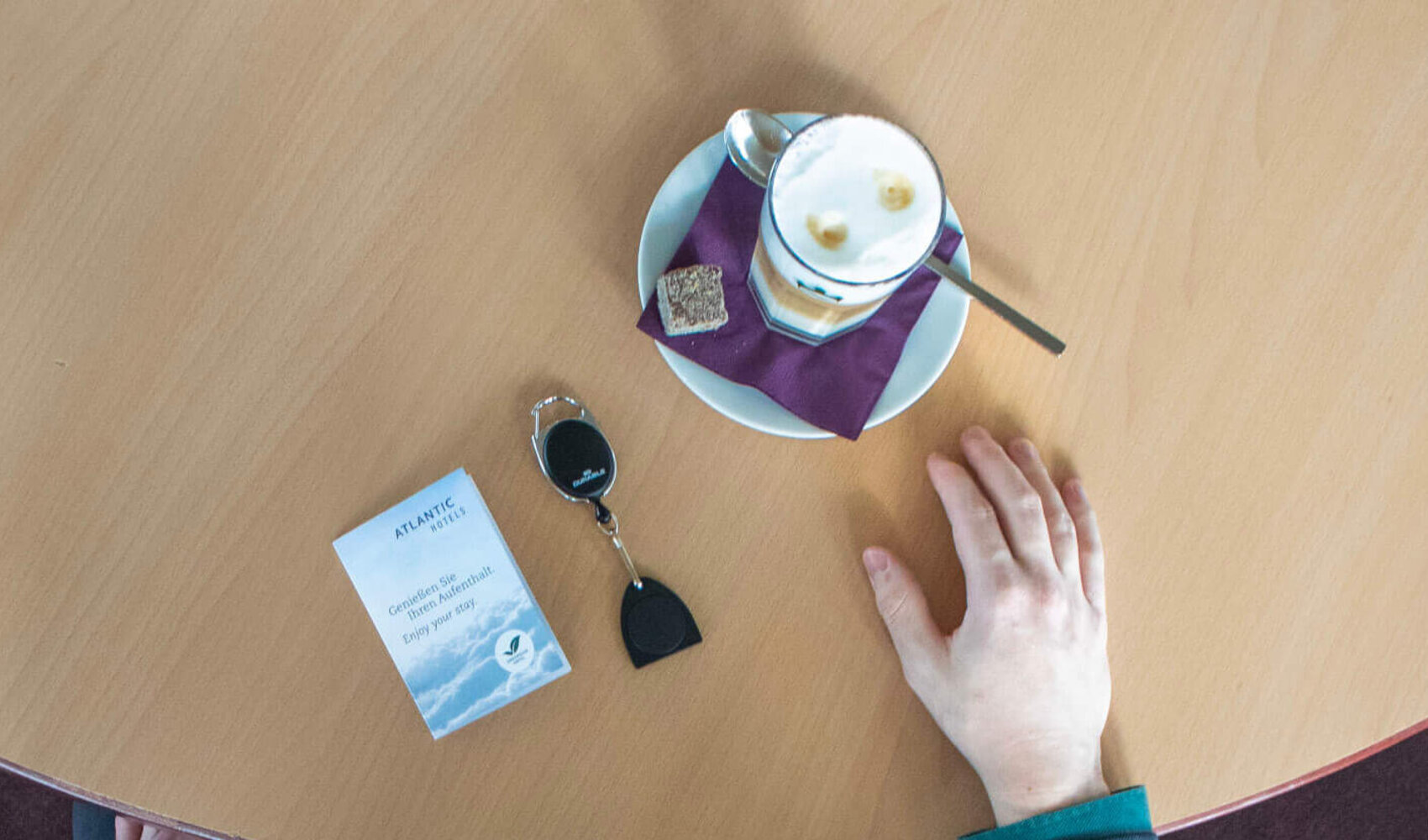Von oben fotografiert ist der dunkelhaarige Florian Ladewig zu sehen wie er an einem hellen Holztisch sitzt. Eine Hand liegt auf dem Tisch, die andere auf seinem Bein. Ein Latte Macchiato steht und eine Schlüsselkarte sowie ein Chip-Schlüssel liegen vor ihm auf dem Tisch. 