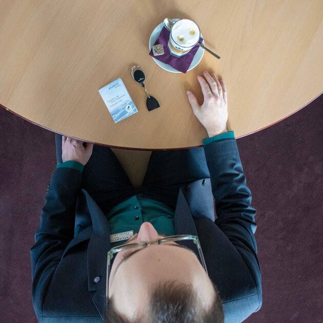 Von oben fotografiert ist der dunkelhaarige Florian Ladewig zu sehen wie er an einem hellen Holztisch sitzt. Eine Hand liegt auf dem Tisch, die andere auf seinem Bein. Ein Latte Macchiato steht und eine Schlüsselkarte sowie ein Chip-Schlüssel liegen vor ihm auf dem Tisch. 