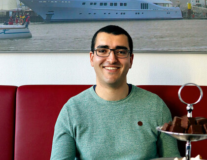 A coffee with temporary helper Mehdi Kebdani Dark-haired temp Mehdi Kebdani sits smiling and wearing glasses in a green sweater on a red upholstered bench in the restaurant. Plates and cups are laid out on the table with a white tablecloth and a large etagere with wintery pastries stands on top. In the background is a picture of a yacht and a sailing boat on a shipyard site.